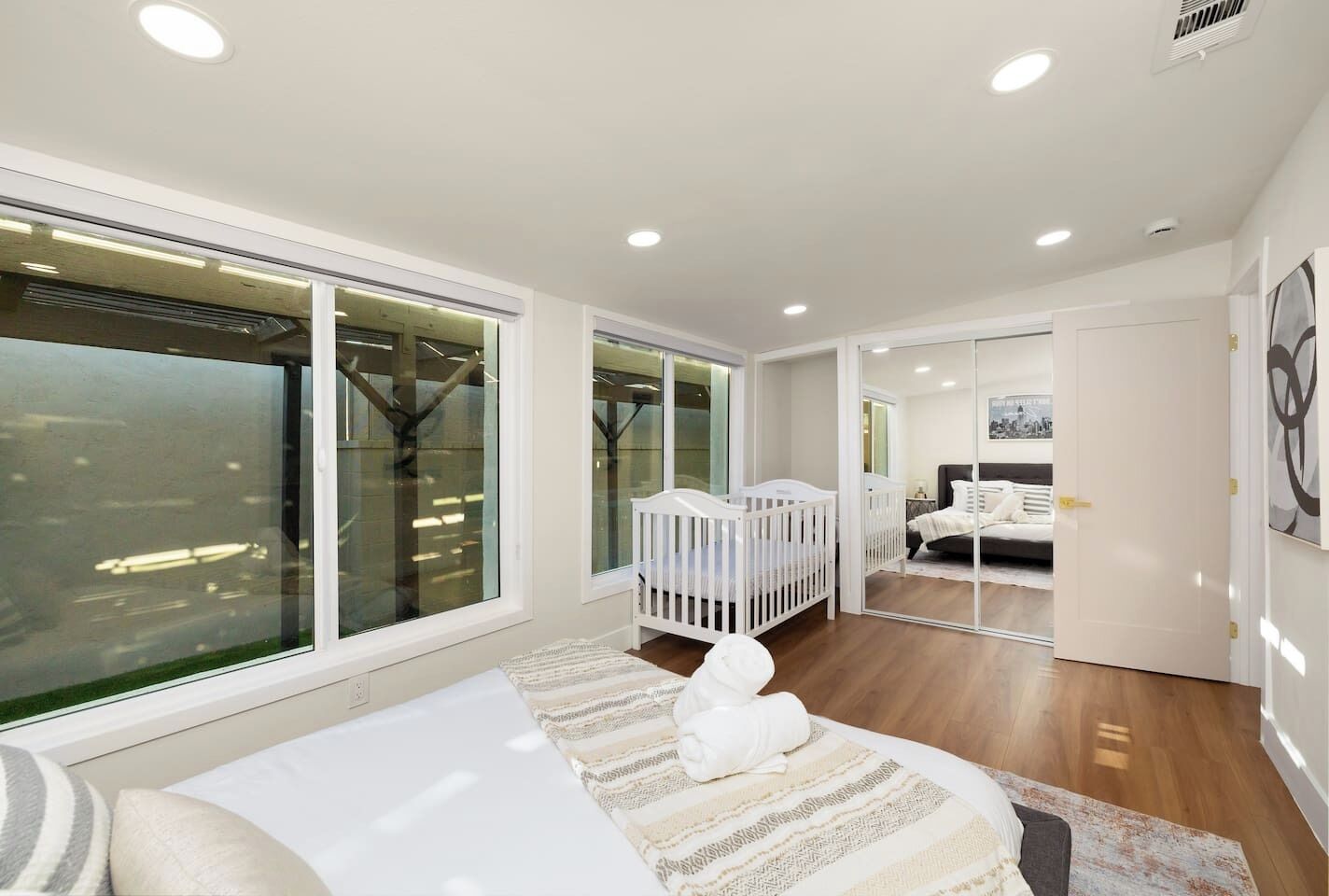 A modern bedroom with a large window, a crib, a bed, and a bathroom visible through a glass door.