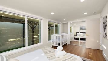 A modern bedroom with a large window, a crib, a bed, and a bathroom visible through a glass door.