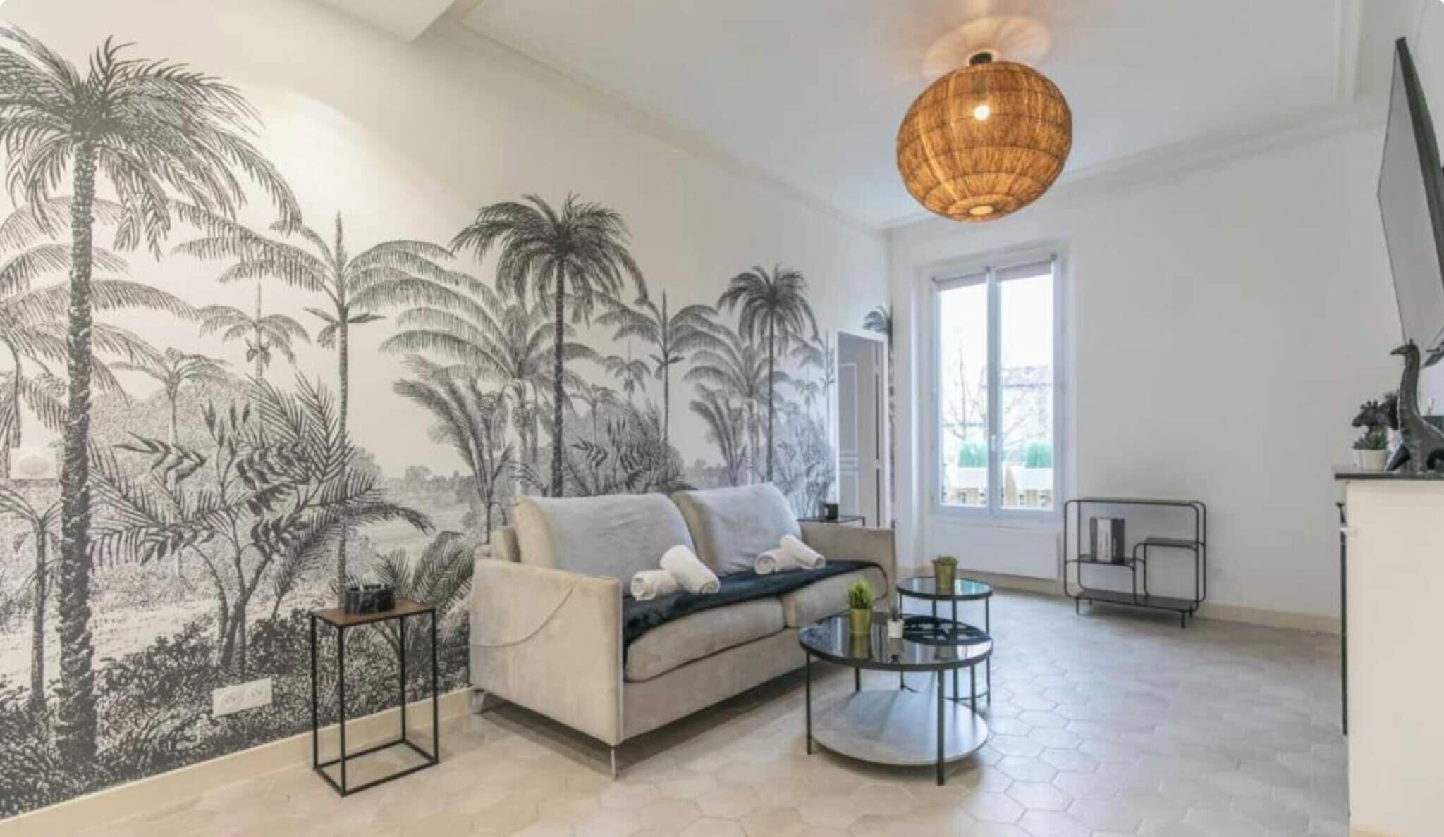 Een woonkamer met een bank, een rond bijzettafeltje, een hanglamp en een muurschildering van palmbomen.