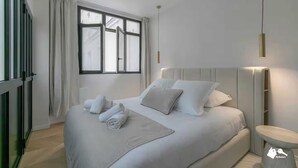 Ett modernt hotellrum med en stor säng, ett sängbord, en liten avlastningsyta och ett fönster med gardiner.