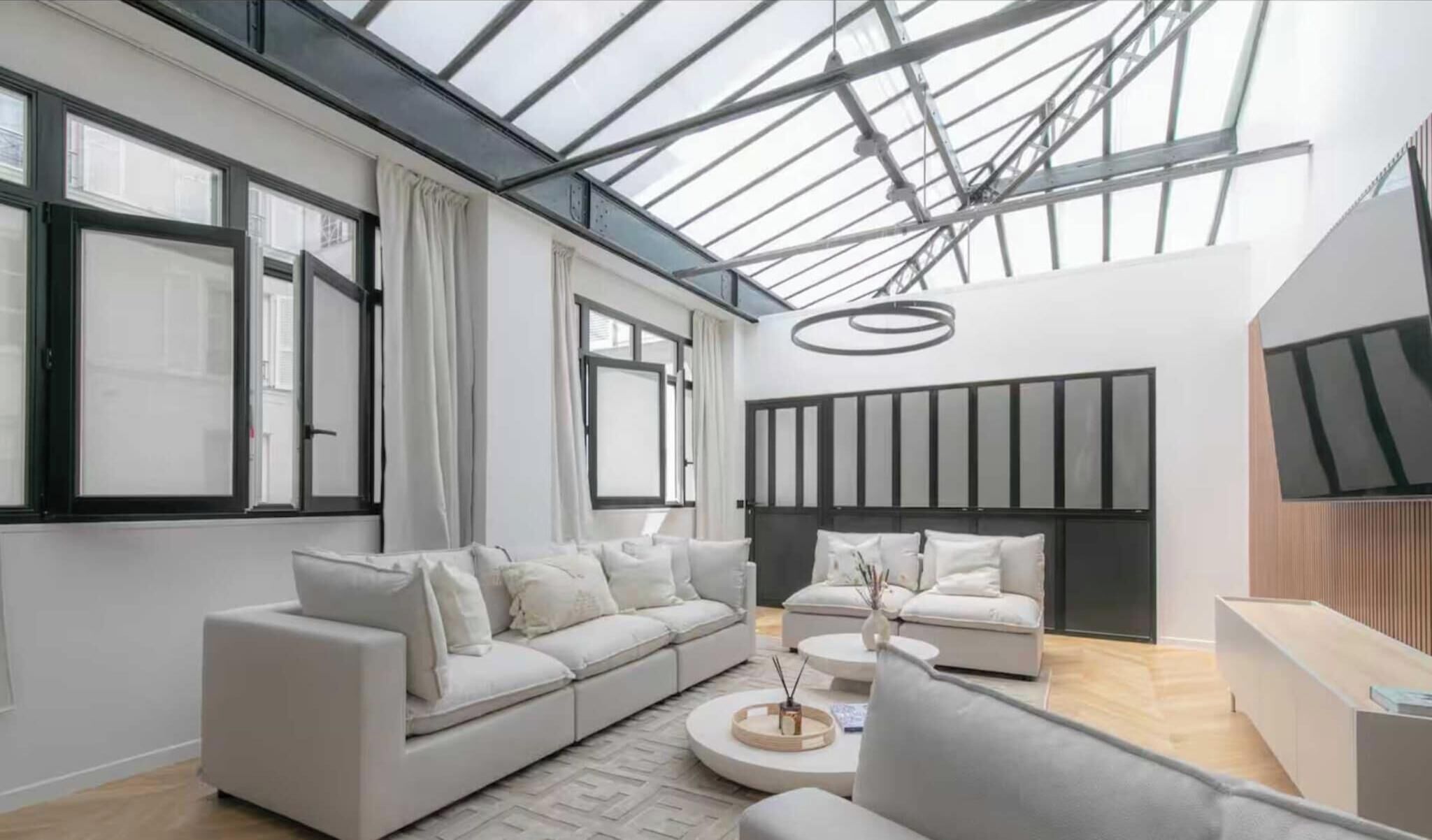 A modern living room with a large sofa, a smaller sofa, and a coffee table. There is a skylight above the sofas, and a flat-screen TV mounted on the wall.