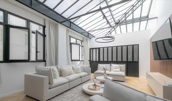 Ein modernes Wohnzimmer mit einer großen Couch, einer kleineren Couch und einem Couchtisch. Über den Sofas befindet sich ein Oberlicht, und an der Wand ist ein Flachbildfernseher angebracht.