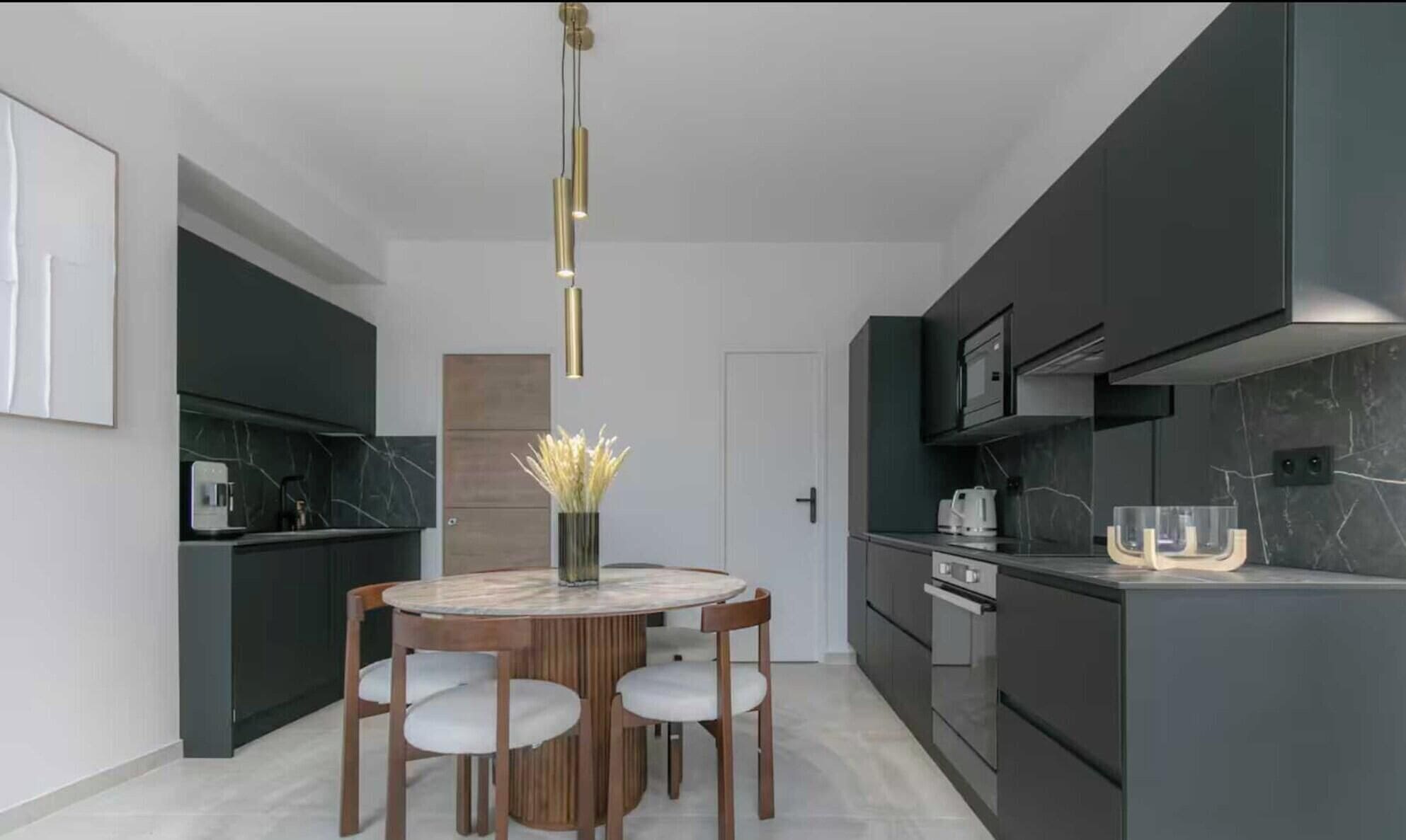 A modern kitchen with dark cabinetry, a marble countertop, and a round dining table with chairs.