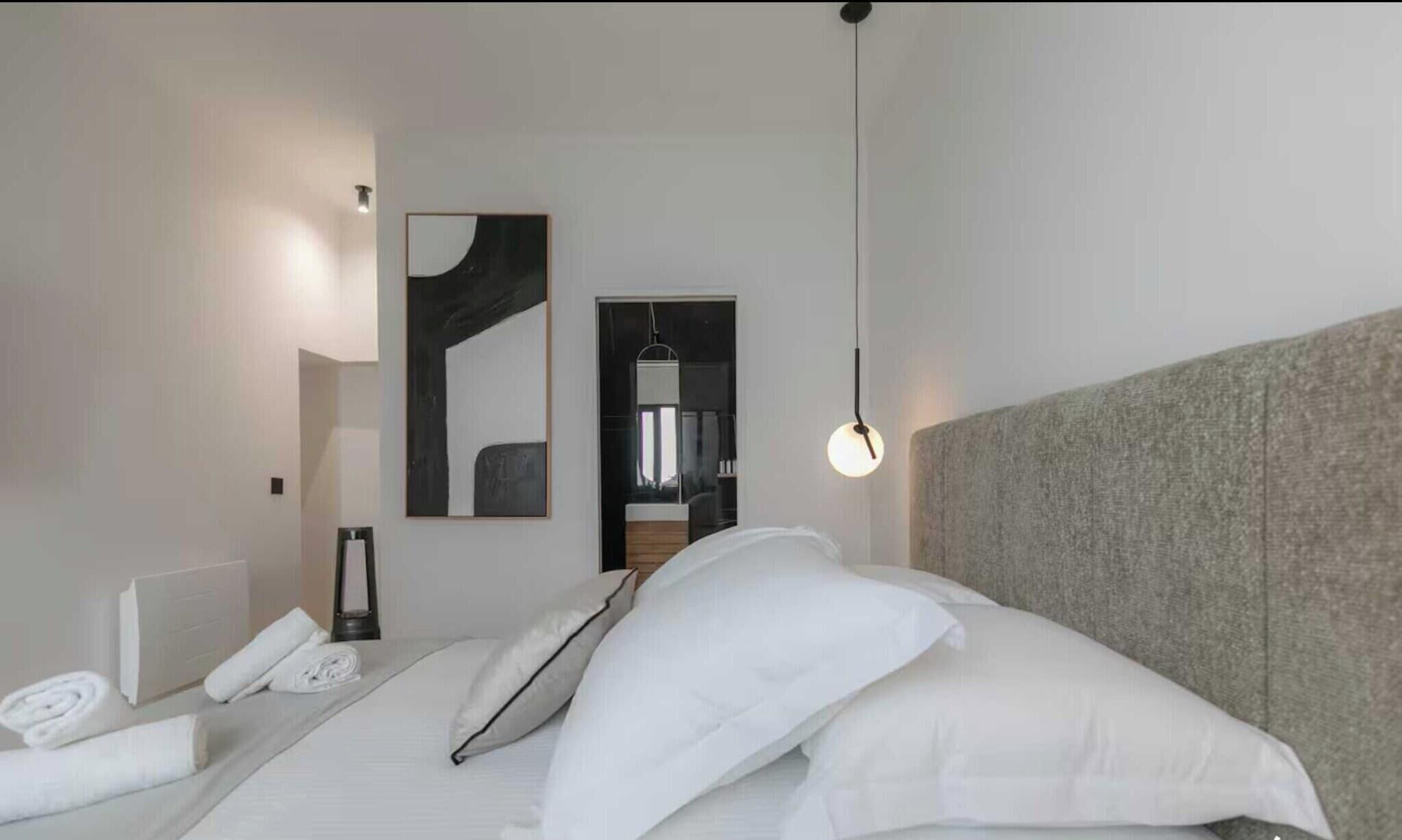 Une chambre d’hôtel moderne avec un lit soigneusement fait, une table de chevet avec une lampe, une grande peinture abstraite au mur et une vue sur la salle de bain par une porte ouverte.