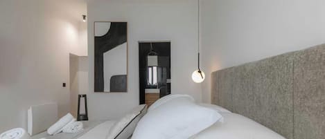 Habitación de hotel moderna con una cama bien hecha, una mesita de noche con lámpara, un cuadro abstracto grande en la pared y vistas al baño a través de una puerta abierta.