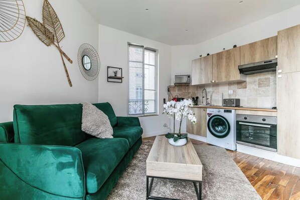 A modern living room with a green sofa, a coffee table, and a kitchen area with wooden cabinets and appliances. - Charming apartment - 2BR/6P - Montreuil near Paris (Montreuil)