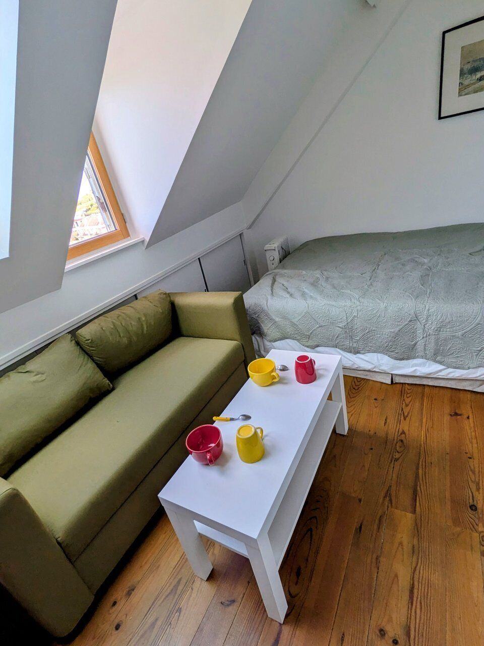Ein kleiner, gemütlicher Raum mit schrägem Dach, einem grünen Sofa, einem weißen Tisch mit Tassen und einem Bett mit einem gemusterten Überwurf.