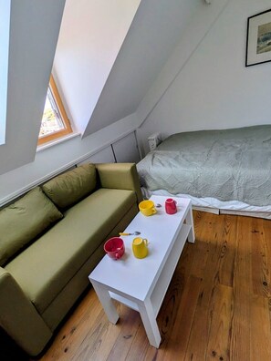 Une petite pièce chaleureuse avec un plafond en pente, un canapé vert, une table blanche avec des tasses et un lit recouvert d’une couverture à motifs.