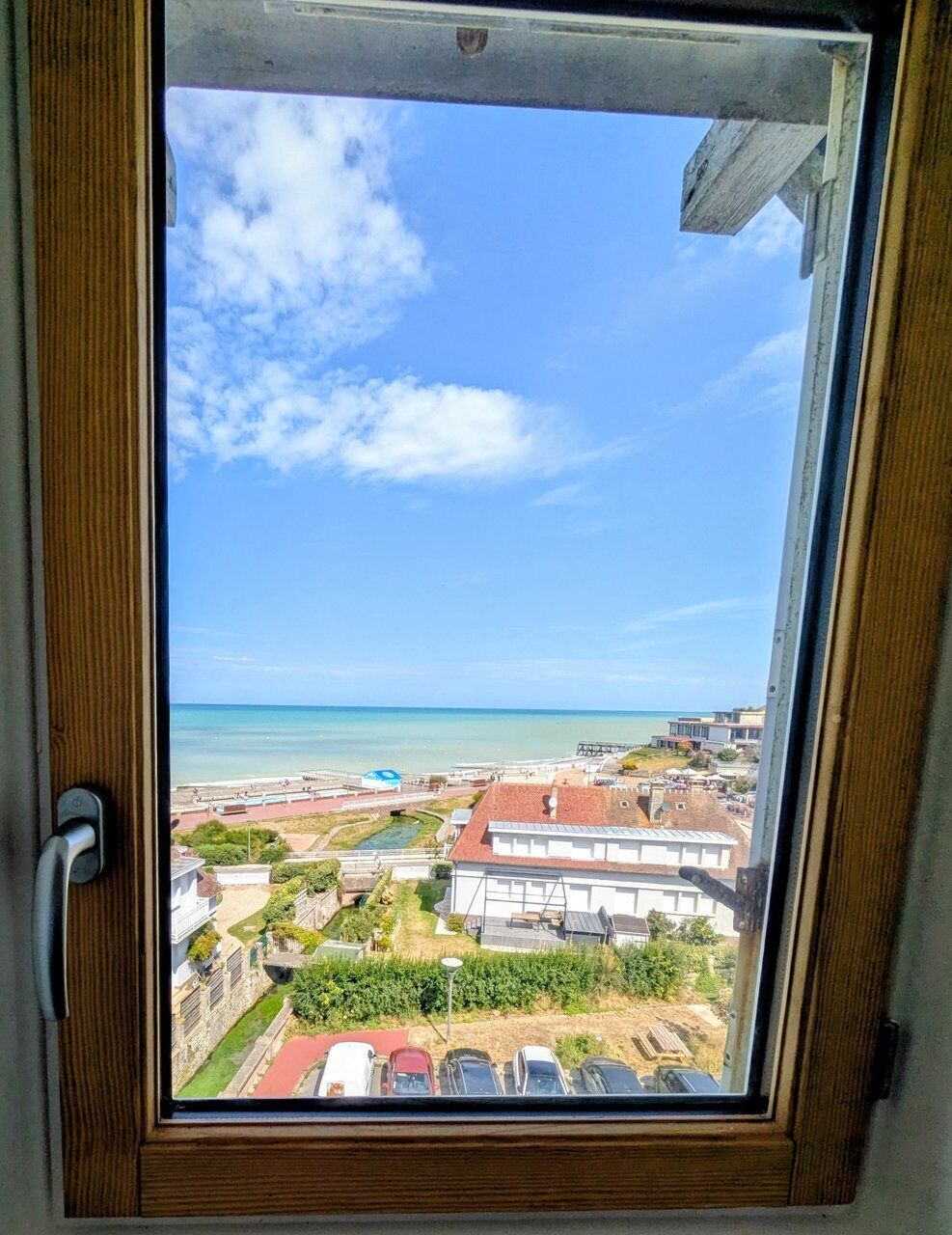 A view through a window with a wooden frame, overlooking a beach, buildings, and a clear sky.