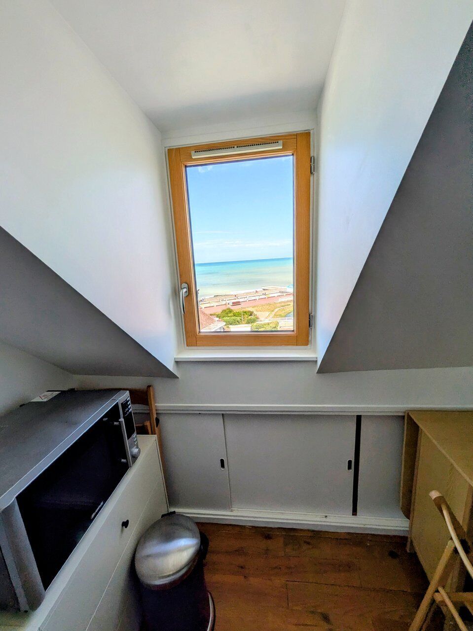 Ein kleiner Dachraum mit geneigter Decke, einem Fenster mit Blick auf den Strand und eingebauten Stauraumschränken.