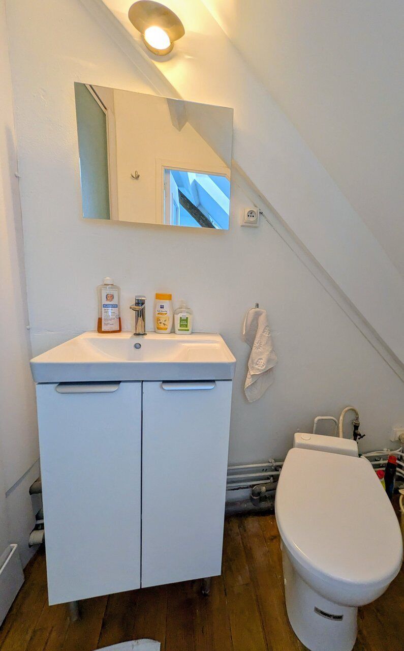 Et badeværelse med en hvid vask, spejl og toilet. Der er toiletartikler på vasken, og et håndklæde hænger på væggen.