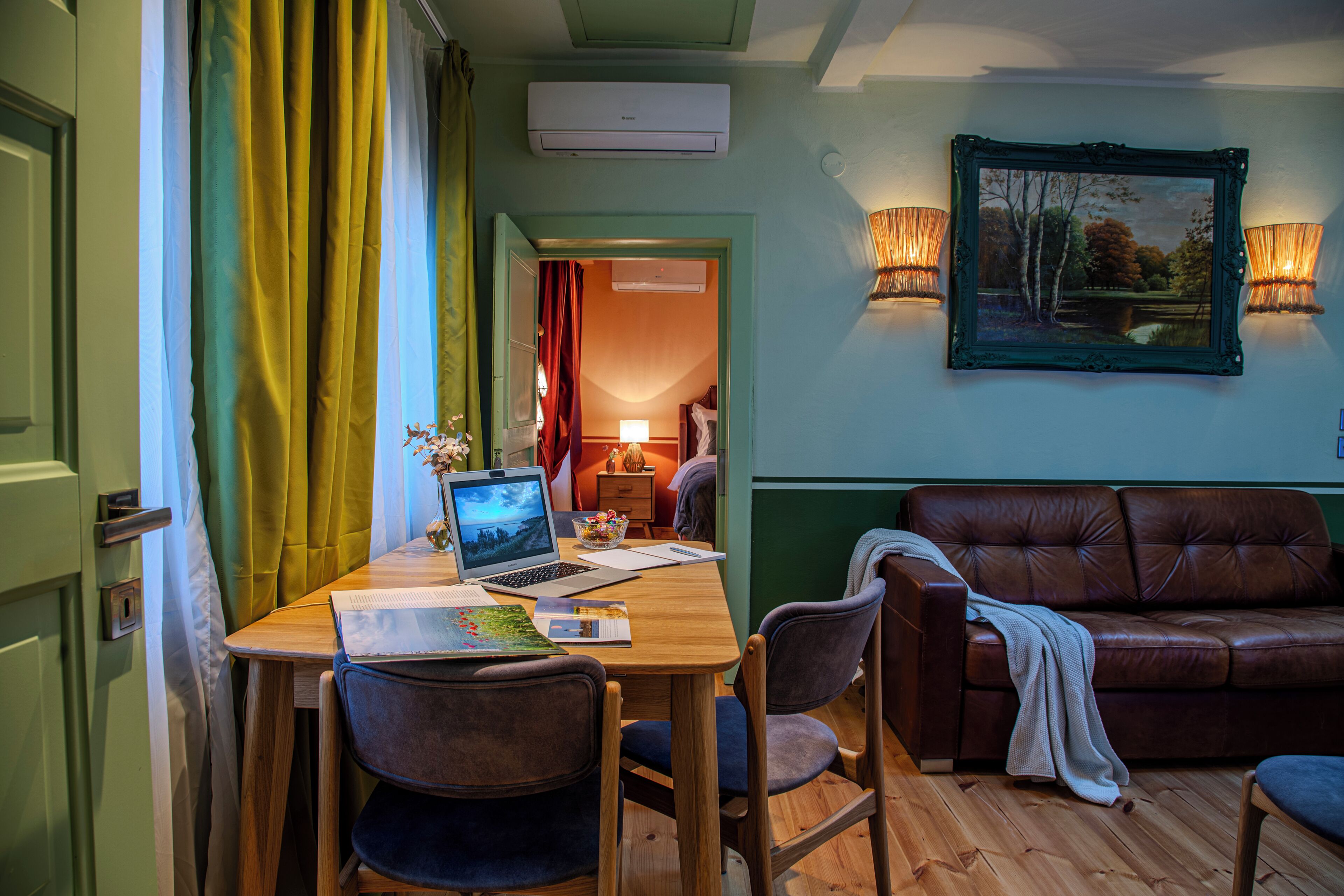 A room with a wooden desk, laptop, chairs, a brown leather sofa, a painting, and a wall-mounted lamp.