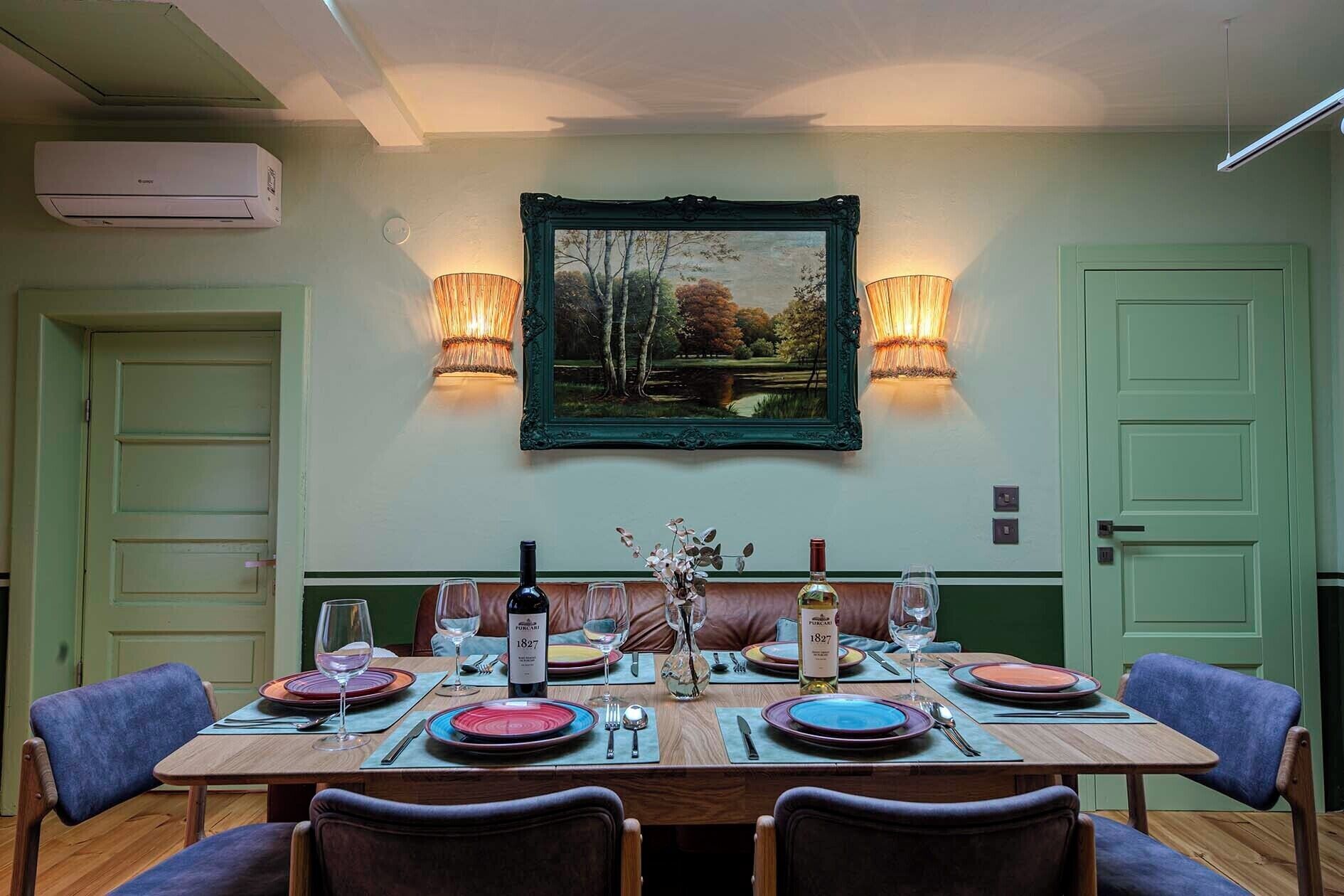 Un coin repas avec une table dressée pour quatre personnes, deux lampes fixées au mur, un tableau encadré et une porte verte.