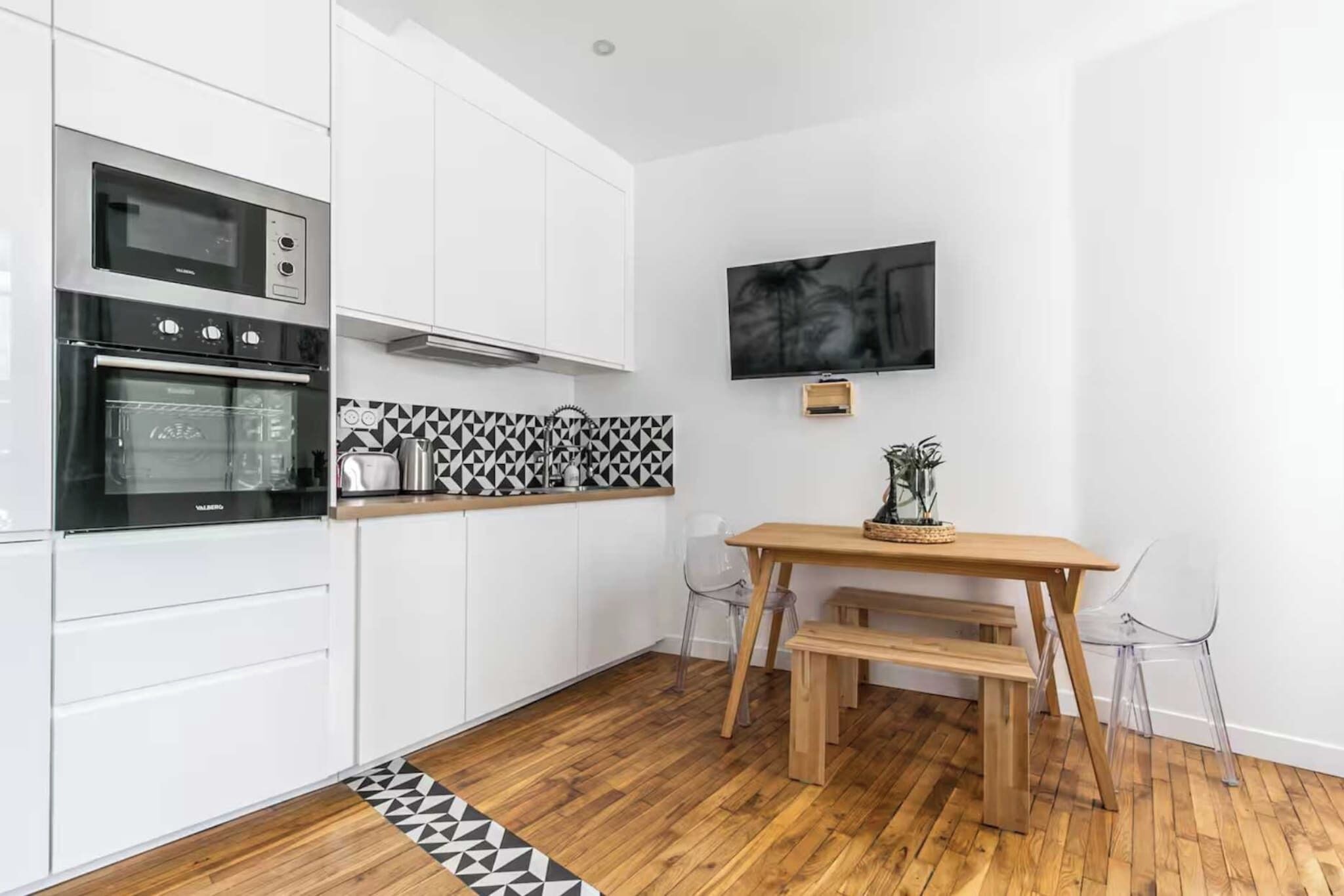 A modern kitchen with white cabinets, a black and white backsplash, a wooden dining table with chairs, and a flat-screen TV mounted on the wall.