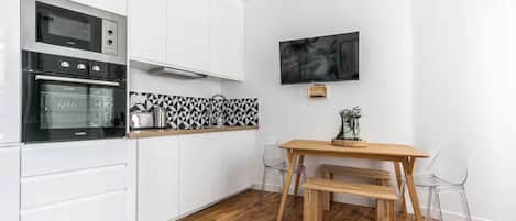 A modern kitchen with white cabinets, a black and white backsplash, a wooden dining table with chairs, and a flat-screen TV mounted on the wall.