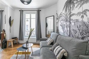 Un salon moderne avec un canapé gris, une petite table ronde et un mur décoratif orné d’une fresque aux motifs tropicaux.
