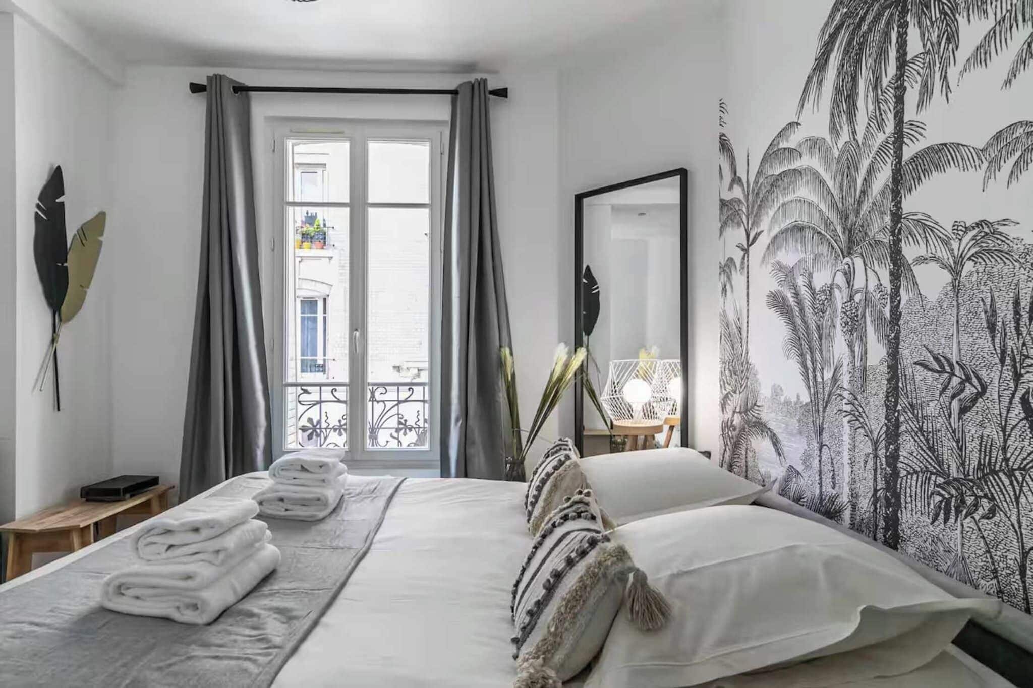A hotel room with a bed, a mirror, a wall with a tropical plant mural, and a balcony with a view.