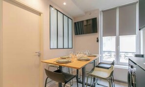 Een eethoek met een tafel voor vier personen, stoelen, een aan de muur bevestigde televisie en een raam met balkon.