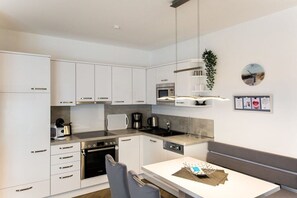 A modern kitchen with white cabinets, stainless steel appliances, and a dining area with a white table and grey chairs. - 7 in Villa Meeresblau (Cuxhaven)