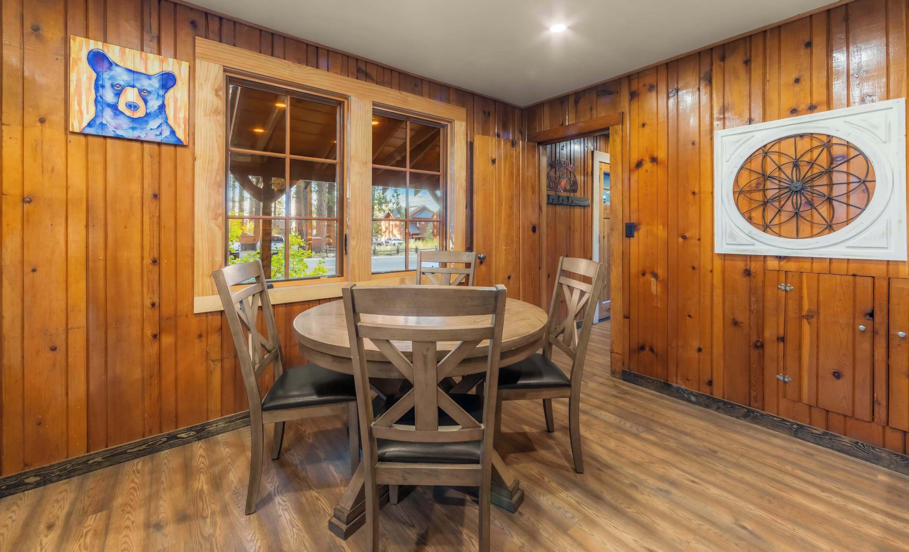 A wooden interior with a round dining table and chairs, a large window with multiple panes, and a bear-themed artwork on the wall.