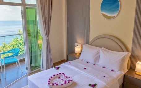 A hotel room with a bed, a nightstand, a lamp, a view of the sea, and a chair by the window.