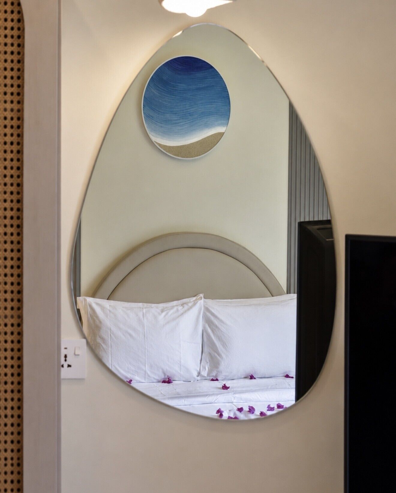 A bed with white linens and purple flower petals, a round mirror on the wall, and a television to the right.