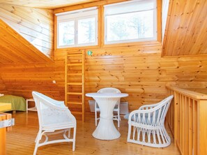 A cozy wooden cabin interior with a loft bedroom, a dining area with white wicker furniture, and a large window providing natural light. - Koli country club b 6 by Interhome (Lieksa)
