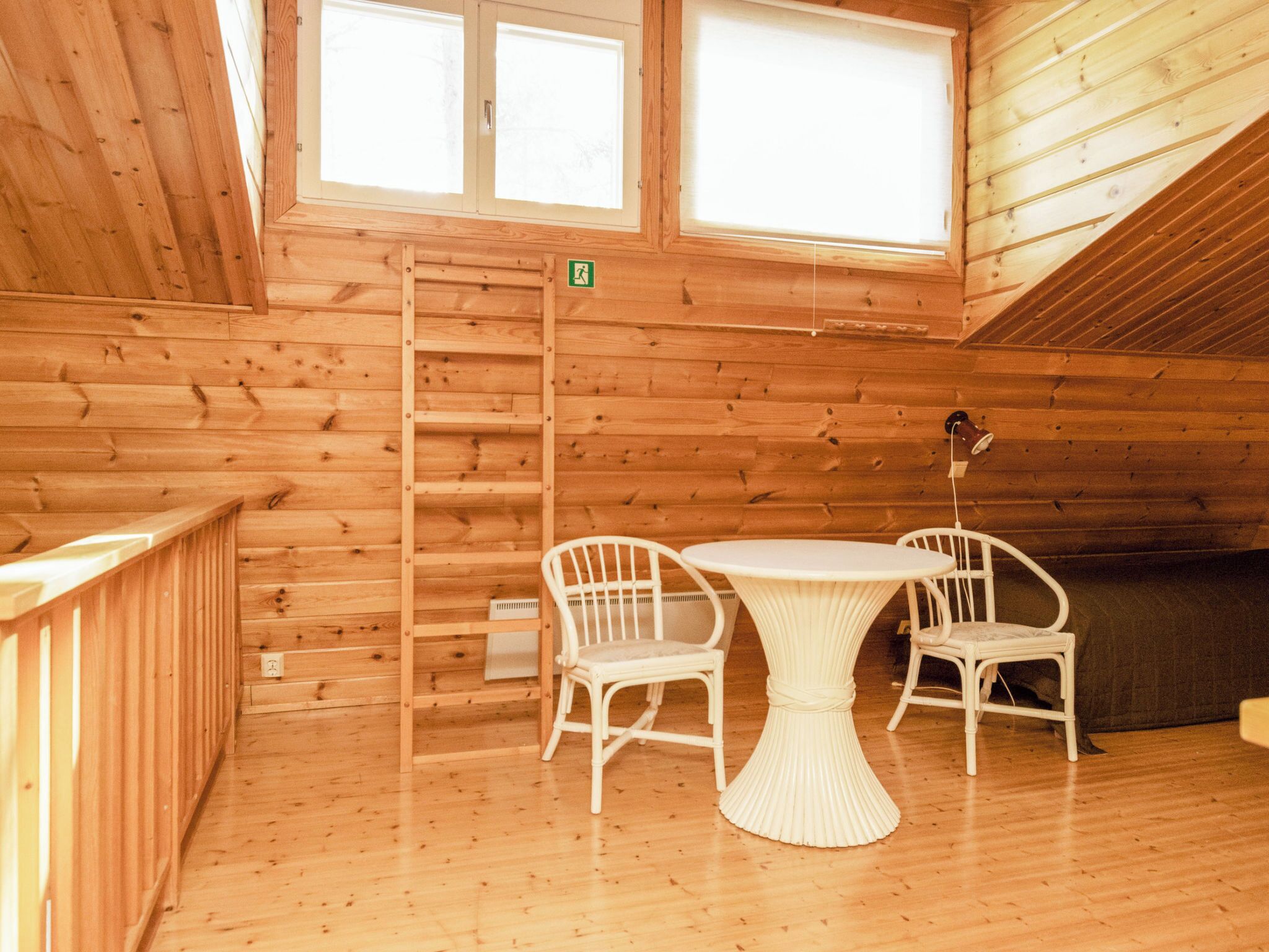 Interior de una cabaña de madera con una mesa de comedor y sillas, una escalera y un ventanal con un letrero de salida verde.