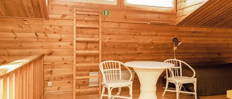 A wooden cabin interior with a dining table and chairs, a ladder, and a window with a green exit sign.