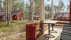 En träveranda med ett bord och stolar, omgiven av en skog med flera röda stugor.
