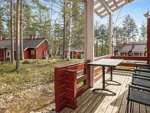 Puinen terassi, jossa on pöytä ja tuoleja, ja sen ympärillä on metsä, jossa on useita punaisia mökkejä.