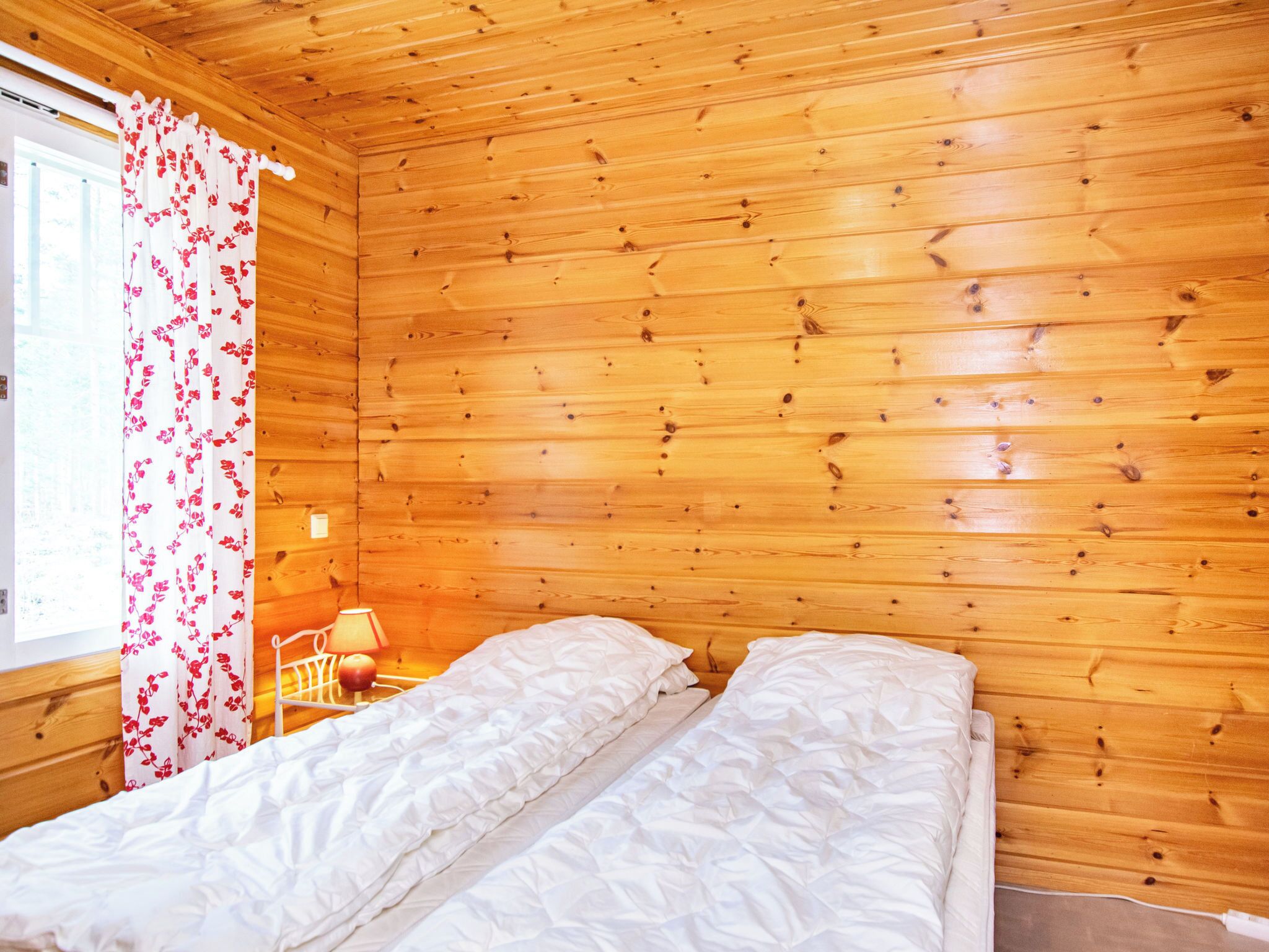 A wooden cabin room with a bed, a nightstand, and a window with curtains.