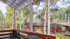 Un espace extérieur abrité, doté d’une table et de chaises, entouré d’une terrasse en bois et offrant une vue sur un bâtiment rouge.