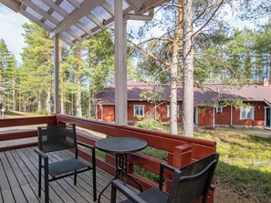 Katettu ulkotila, jossa on pöytä ja tuoleja, ja jota ympäröi puinen terassi. Sieltä on näkymä punaiseen rakennukseen.