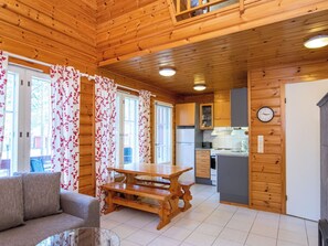 Interior de uma cabana de madeira, com mesa de jantar, sofá, cozinha e janelas amplas com cortinas florais.
