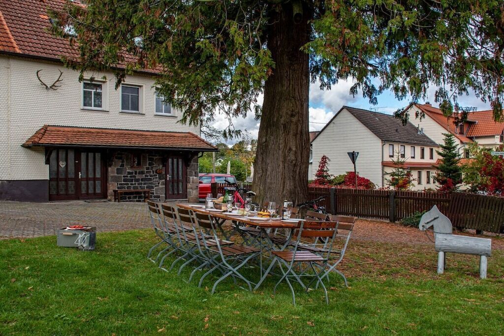 Un espace extérieur aménagé pour les repas, avec une table dressée pour quatre personnes, entourée de chaises en métal, devant un bâtiment à la façade en pierre et un grand arbre.