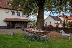 An outdoor dining area with a table set for four, surrounded by metal chairs, in front of a building with a stone facade and a large tree. - Old Forester's Lodge Hella (Grebenhain)