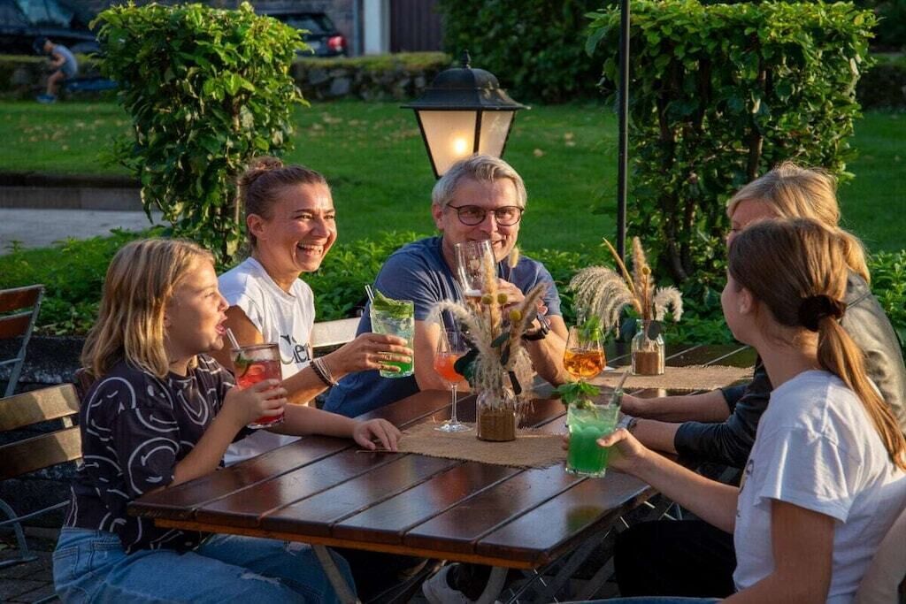 Eine Gruppe von Personen sitzt an einem Tisch im Freien mit Getränken, umgeben von Pflanzen und einem Lampenpfosten.