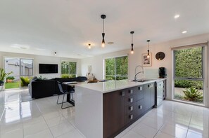 A modern kitchen with a central island, a dining area with a black sofa, and large windows offering a view of greenery.