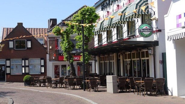 A street with outdoor seating, a building with a Heineken sign, and a tree in the foreground.