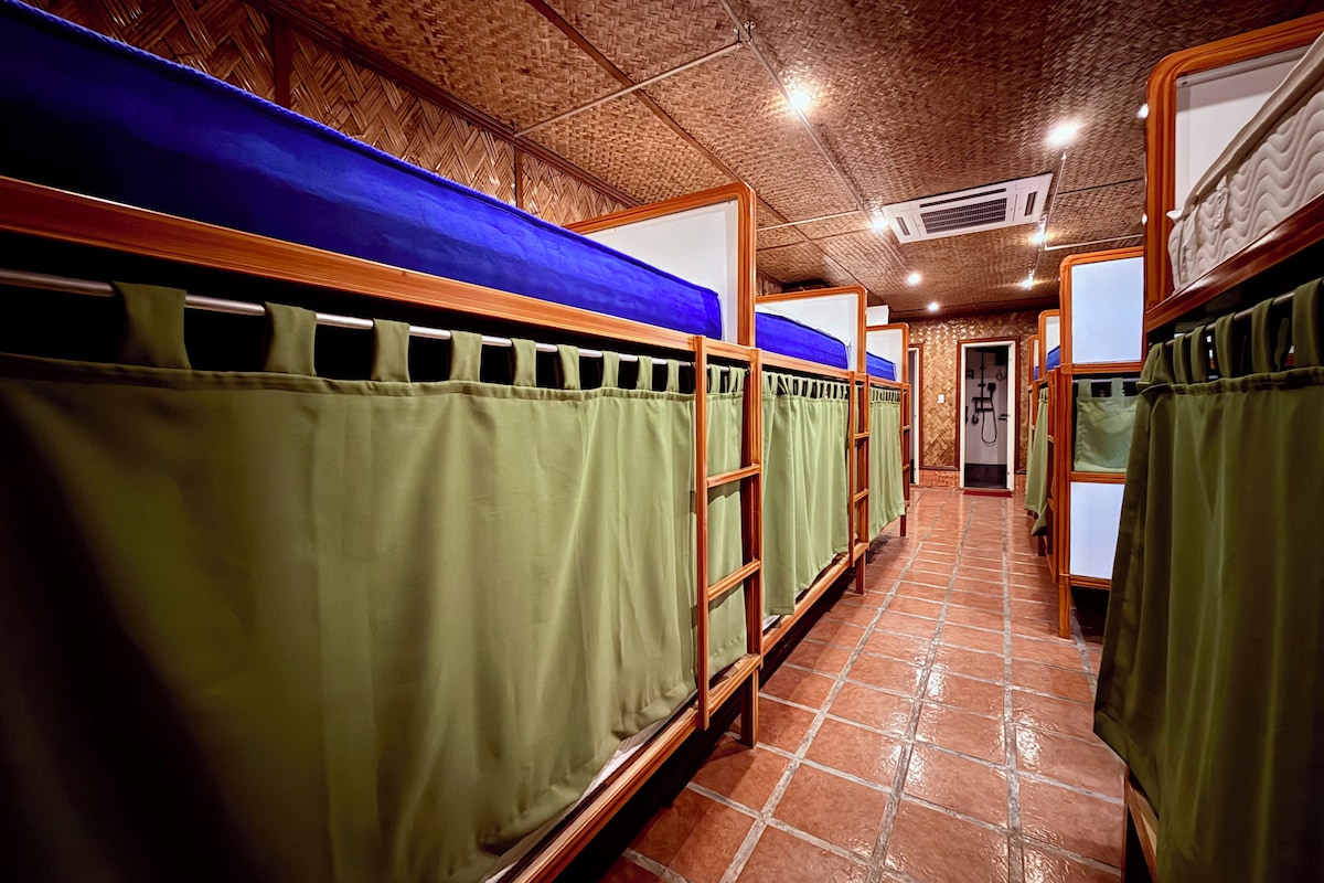 Una hilera de literas con cortinas verdes, armazones de madera y piso de baldosas.