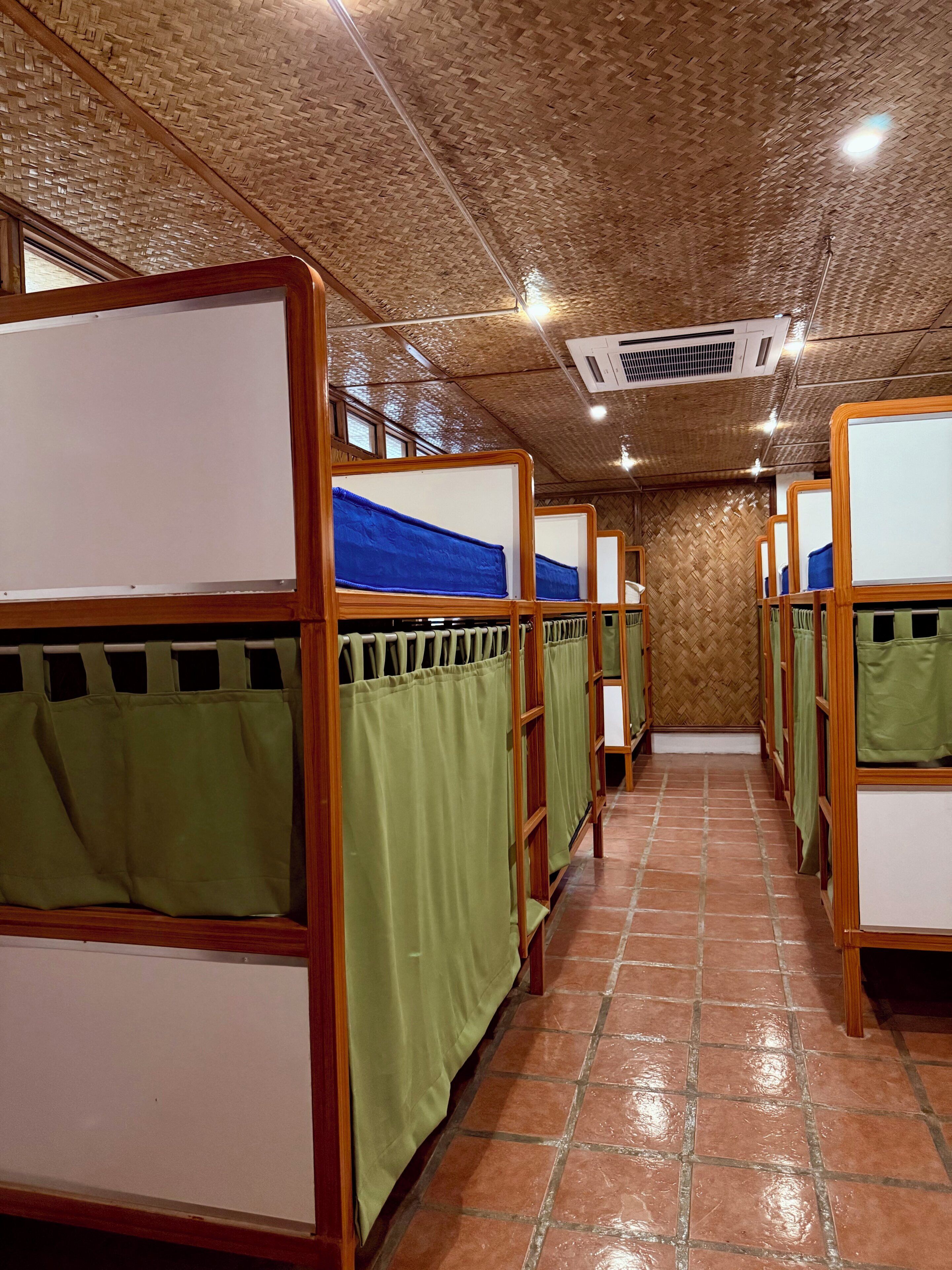 Una fila de literas con cortinas verdes, armazones de madera y techo tejido.