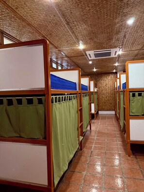 Una fila de literas con cortinas verdes, armazones de madera y techo tejido.