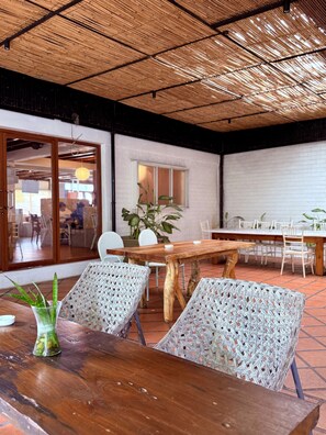 An indoor dining area with a wooden table and woven chairs, a potted plant, and a view of the outside through a glass door.