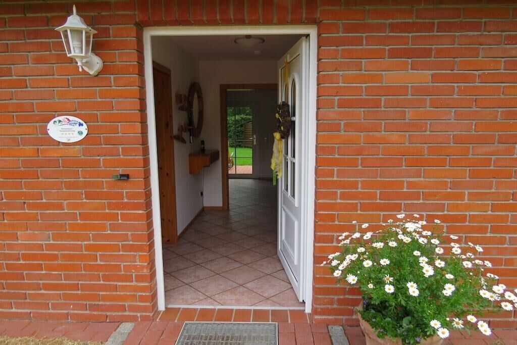A brick building entrance with a white door, a potted plant with white flowers, and a wall-mounted light fixture.