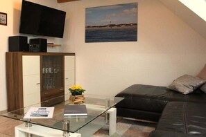 A living room with a glass coffee table, a black leather sofa, a flat-screen TV, and a painting of a beach scene. - HOUSE LUV 2 - on the upper floor (Wittdün)