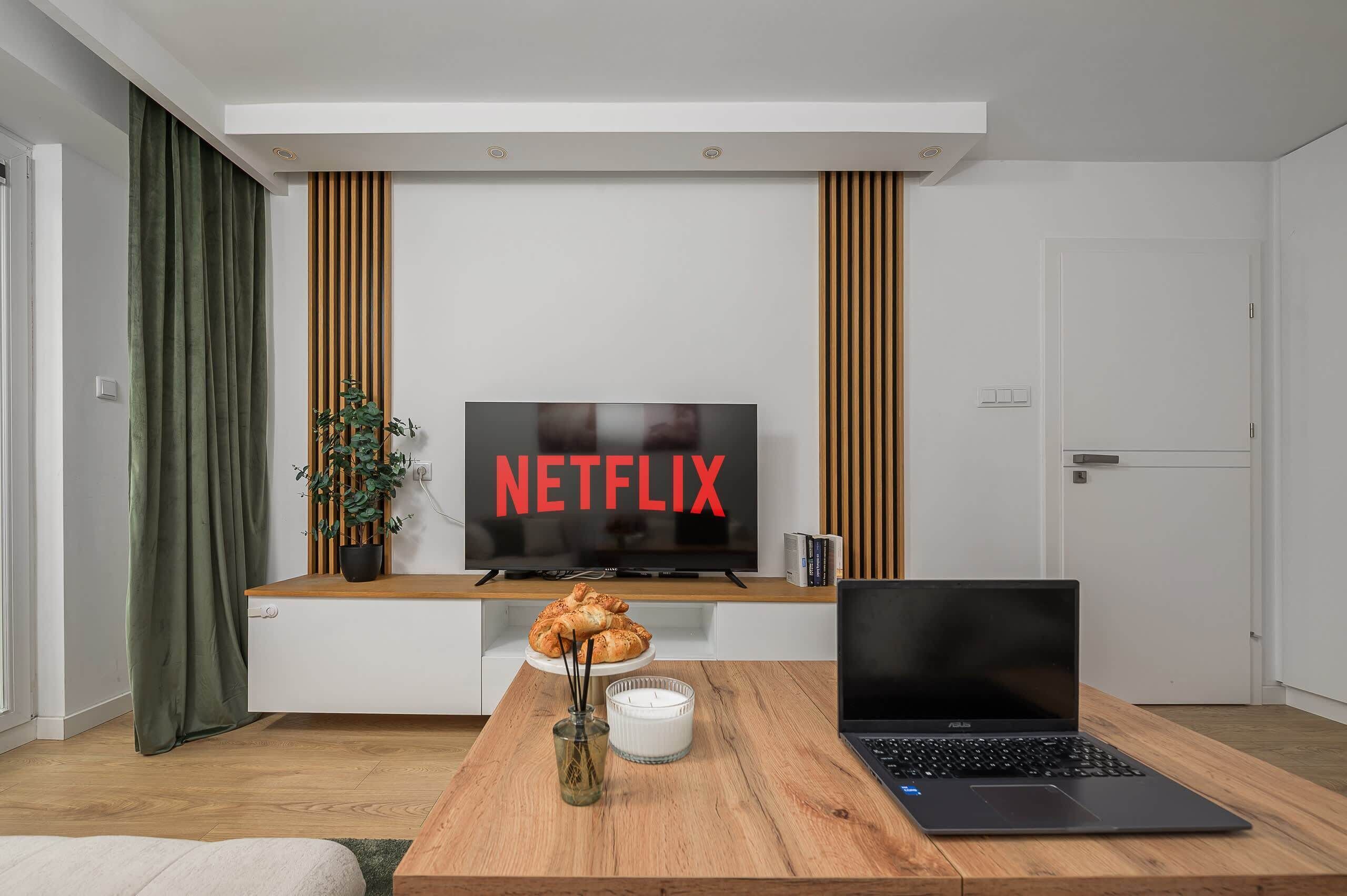 Un salon moderne avec un téléviseur à écran plat affichant le logo « NETFLIX », une table basse en bois sur laquelle sont posés une bougie et un ordinateur portable, et une plante en pot.