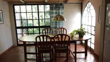 Uma área de refeições com mesa redonda, quatro cadeiras de madeira, luminária pendente e um vaso com planta.