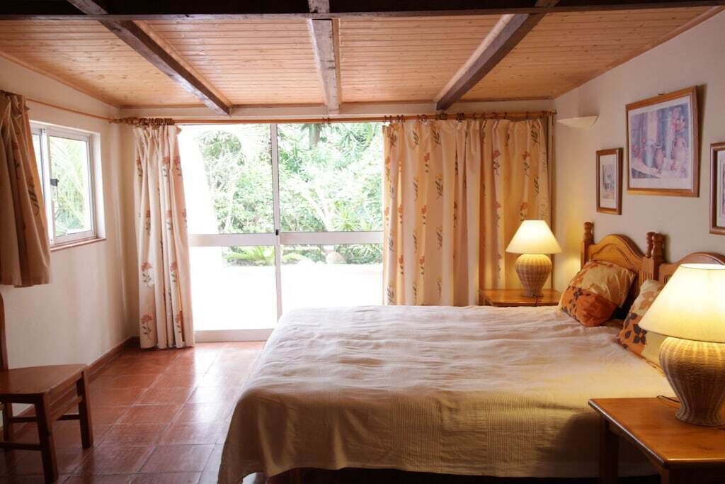 Quarto com cama, duas luminárias laterais, cabeceira de madeira, janela com cortinas e uma porta.