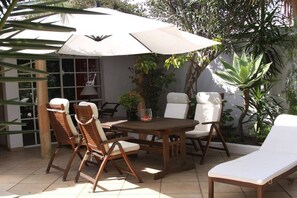 Eine Terrasse im Freien mit einem runden Holztisch, vier Stühlen und einem großen Sonnenschirm, der Schatten spendet.
